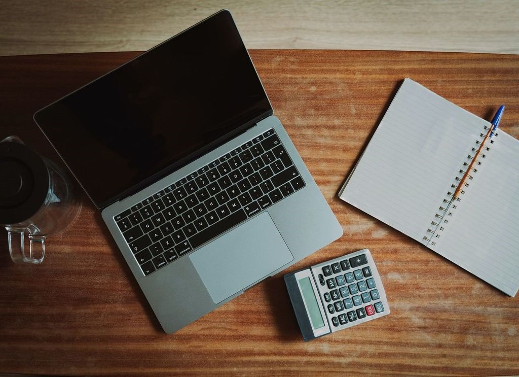 Laptop, calculator, and notebook on a wooden desk, representing an office setup for surveyors and admin personnel